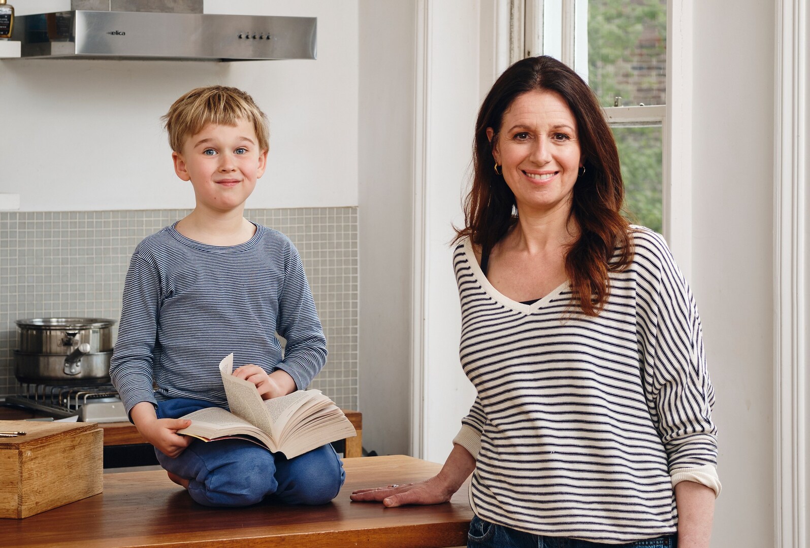 A woman and a young boy sit at a kitchen counter; the boy is reading a book.