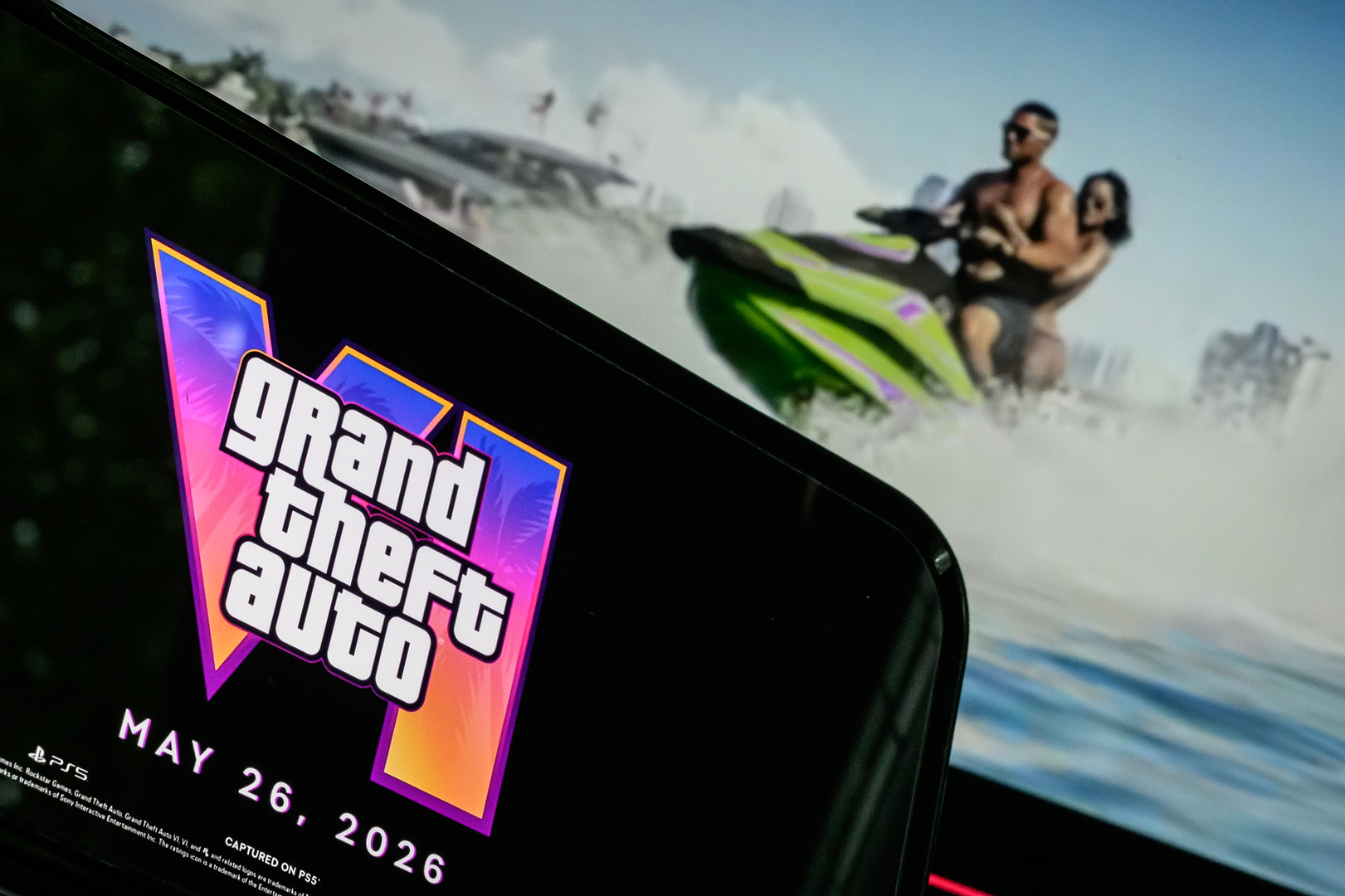 Photo illustration of a phone displaying the Grand Theft Auto VI logo and release date in front of a screen showing a trailer for the game.